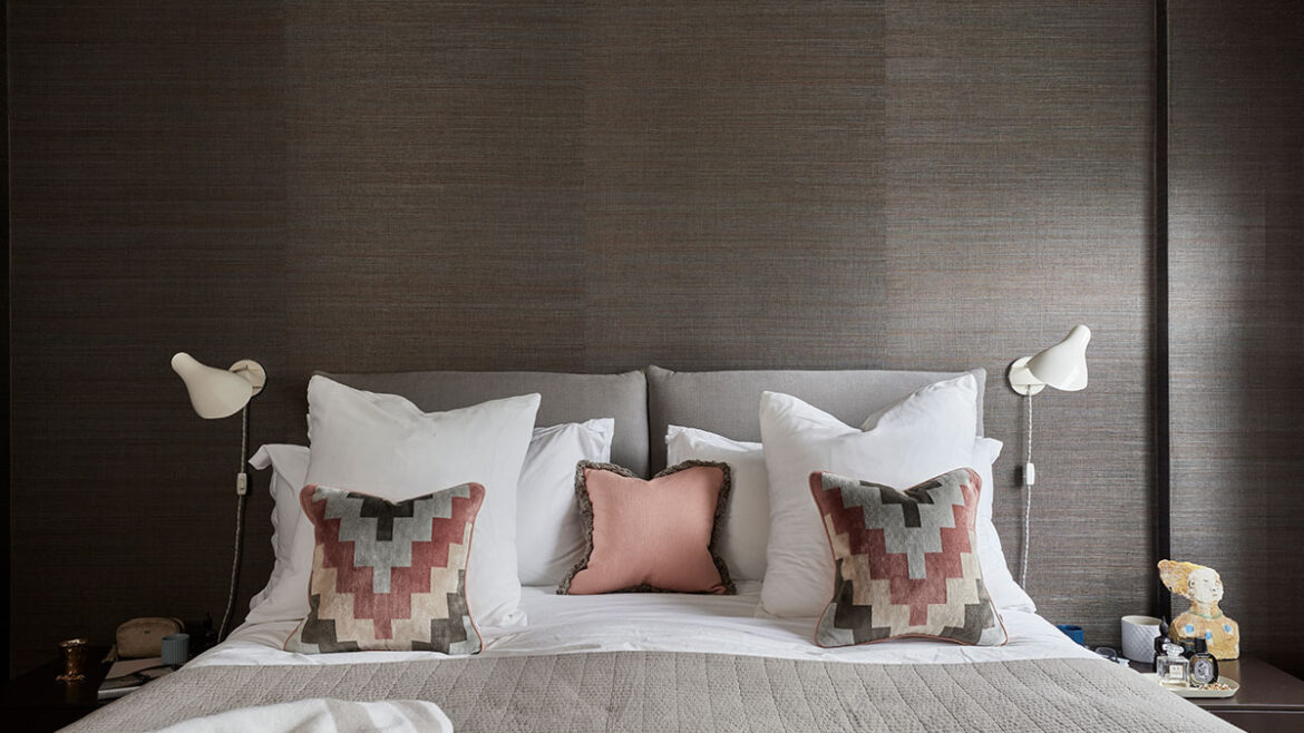 Cosy bedroom with layered pillows in earthy tones against a chocolate brown textured wallpaper backdrop.