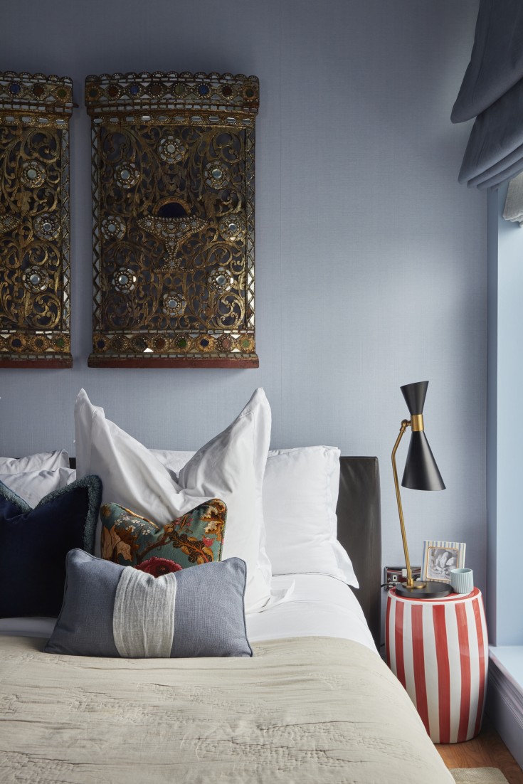 Guest bedroom design in luxury London penthouse with powder blue walls, antique carved panels, striped red and white bedside table, and layered cushions.